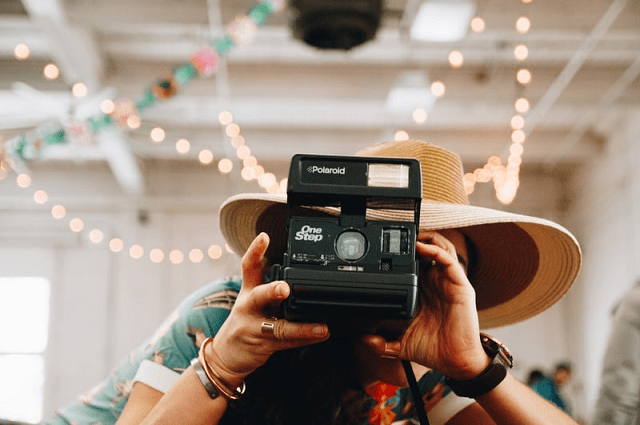 10 consejos para organizar una despedida de soltera de forma virtual,a person wearing a summer shirt and sun har taking a picture with a Polaroid camera in a room decorated with streamers and twinkle lights.